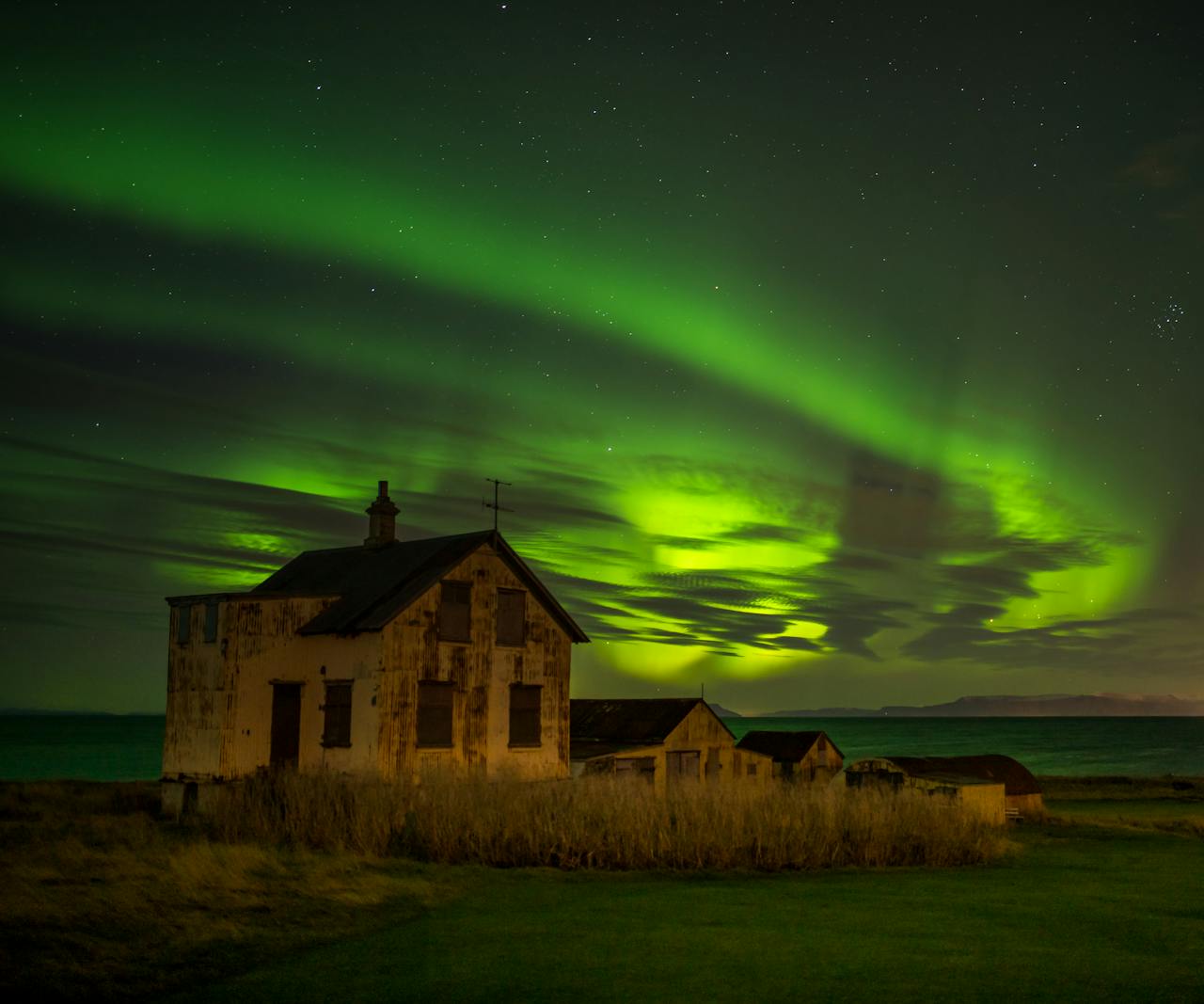 Northern lights illuminate an abandoned house on the coastline of Keflavík, Iceland under a starry night sky.